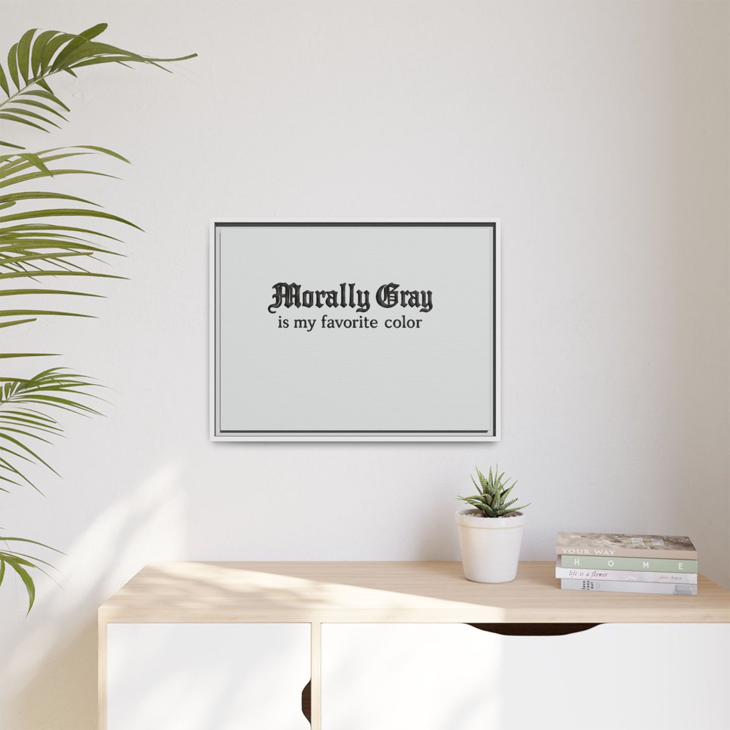 A minimalist framed print on a white wall with the phrase "Morally Gray is my favorite color," displayed above a wooden table holding a small potted plant and a stack of books featuring the Unknown Product.