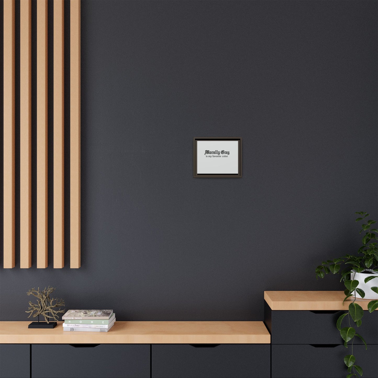 Minimalist modern interior wall with dark gray paint, light wood panel accents, a small framed "Unknown Product" print, and a black cabinet featuring decorative plants and stacked books.
