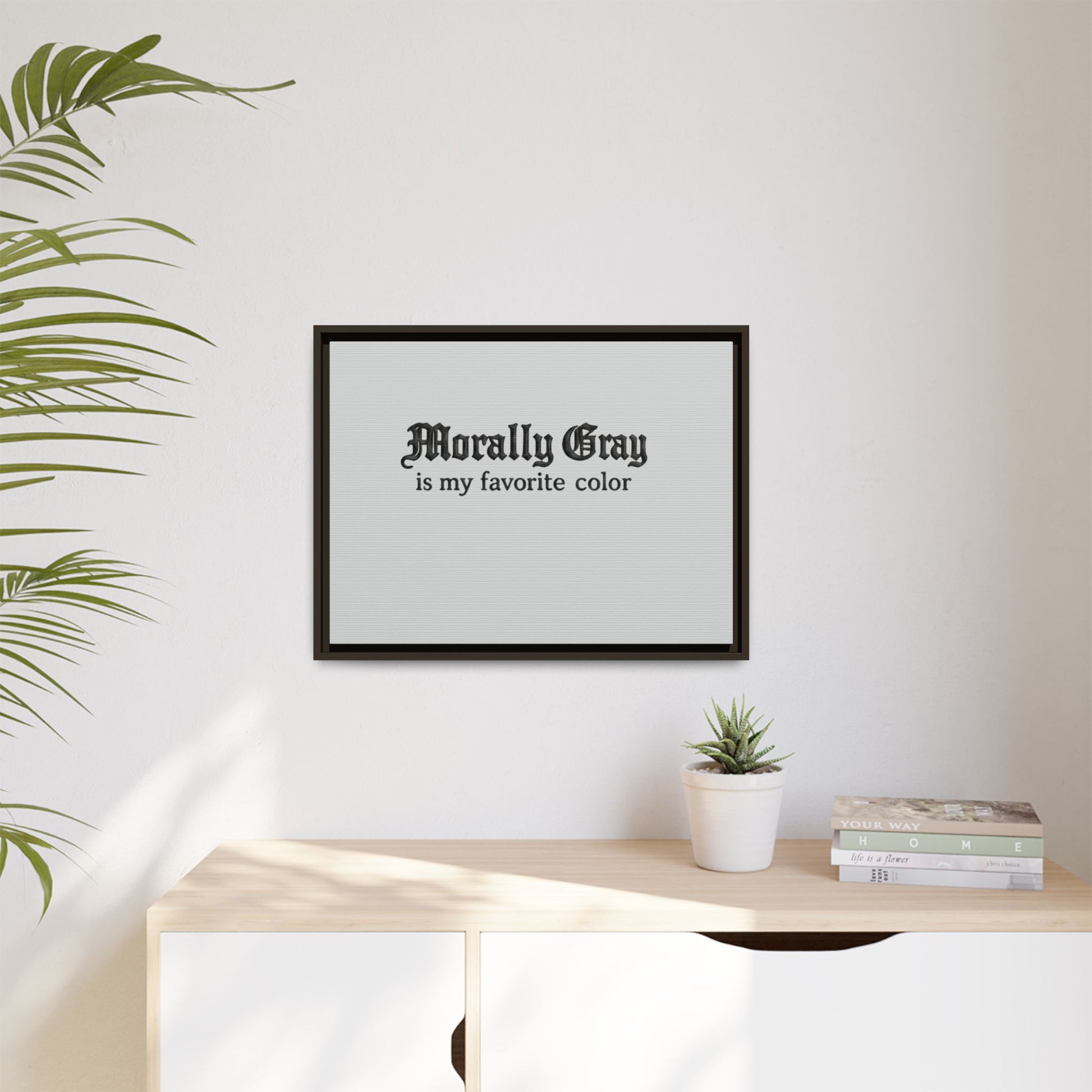 Framed wall art with the phrase "Morally Gray is my favorite color" in black gothic font above a light wooden cabinet, styled with a small potted plant and stacked books, showcasing Unknown Product.