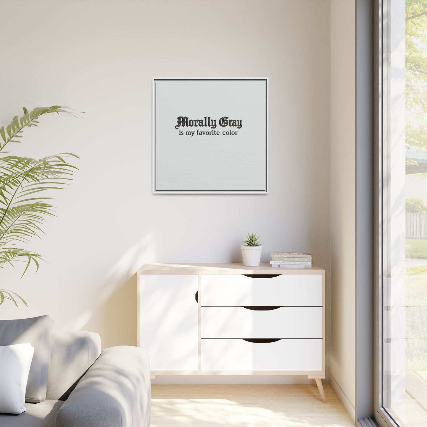 Minimalist living room corner with modern white cabinet, potted plants, and a framed print on the wall reading "Morally Gray is my favorite color," featuring Unknown Product branding.