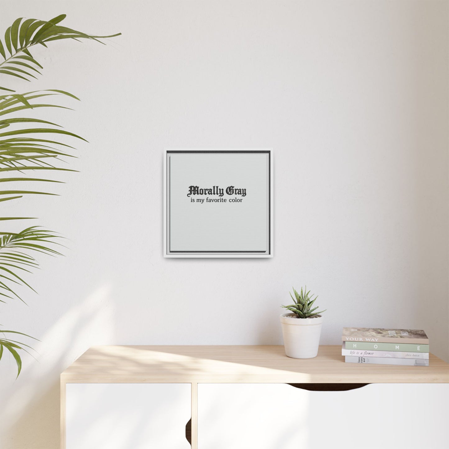 Minimalist wall art featuring the phrase "Morally Gray is my favorite color" in black text displayed above a light wood cabinet with a small potted plant and stacked books, showcasing the Unknown Product.