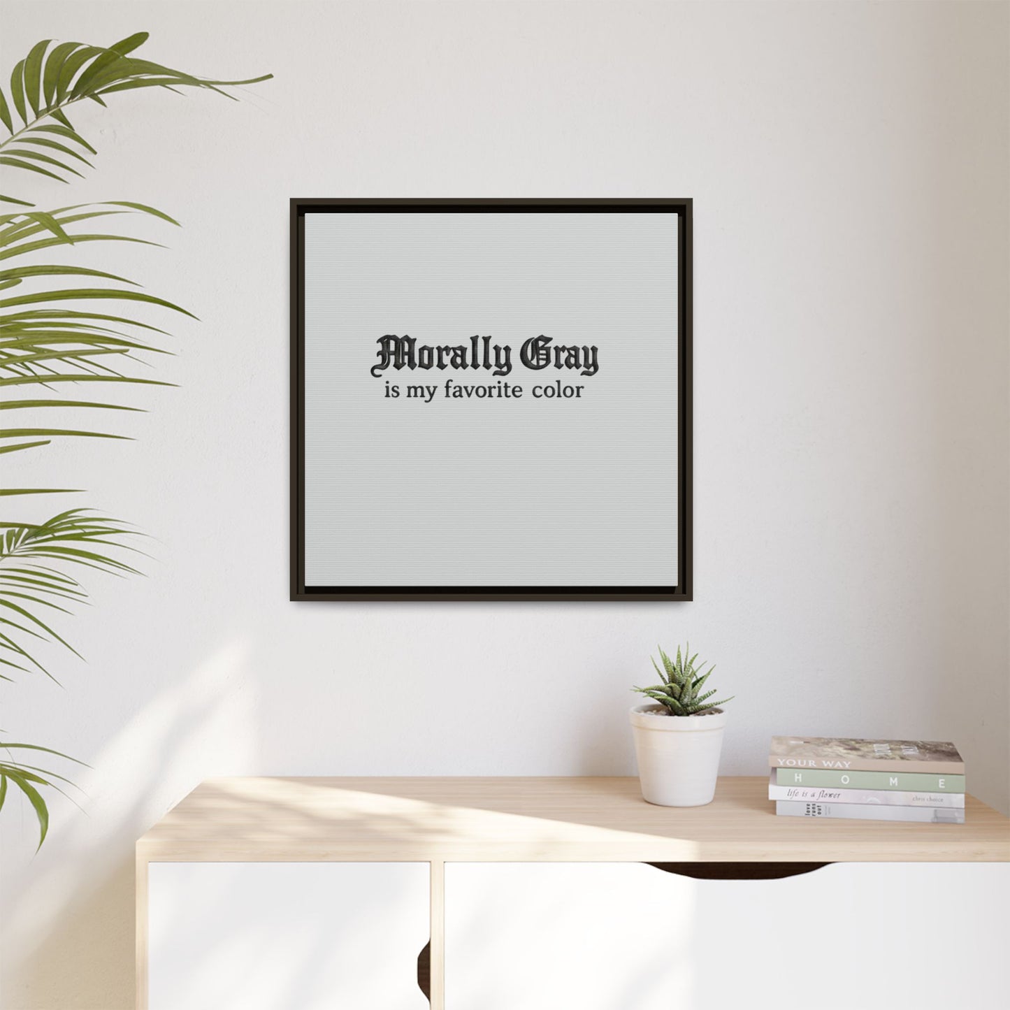 Framed wall art with gothic text reading "Morally Gray is my favorite color" hanging above a wooden dresser with a small potted succulent and stacked books, styled in a minimalist room featuring the Unknown Product.