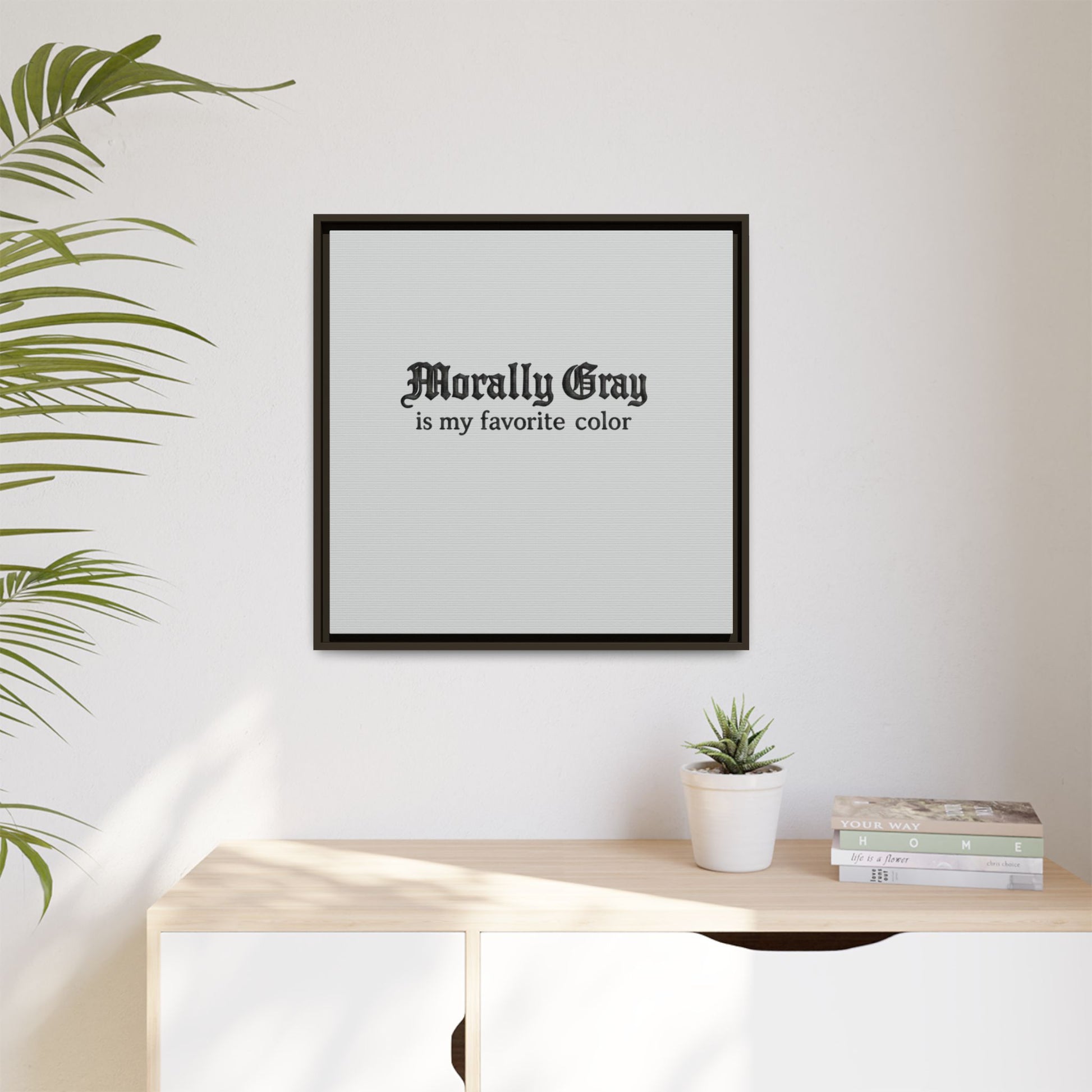 Framed wall art with gothic text reading "Morally Gray is my favorite color" hanging above a wooden dresser with a small potted succulent and stacked books, styled in a minimalist room featuring the Unknown Product.