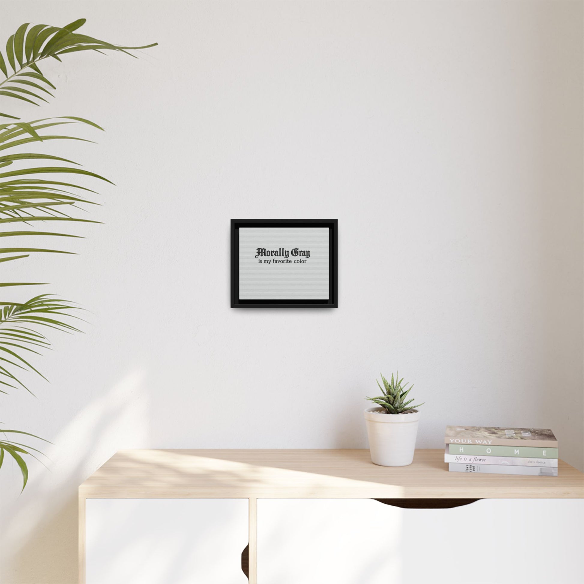 Minimalist framed print with the phrase "Morally Gray - a ray between white" hanging on a white wall above a light wood cabinet with a small potted succulent and stacked books, featuring the Unknown Product.