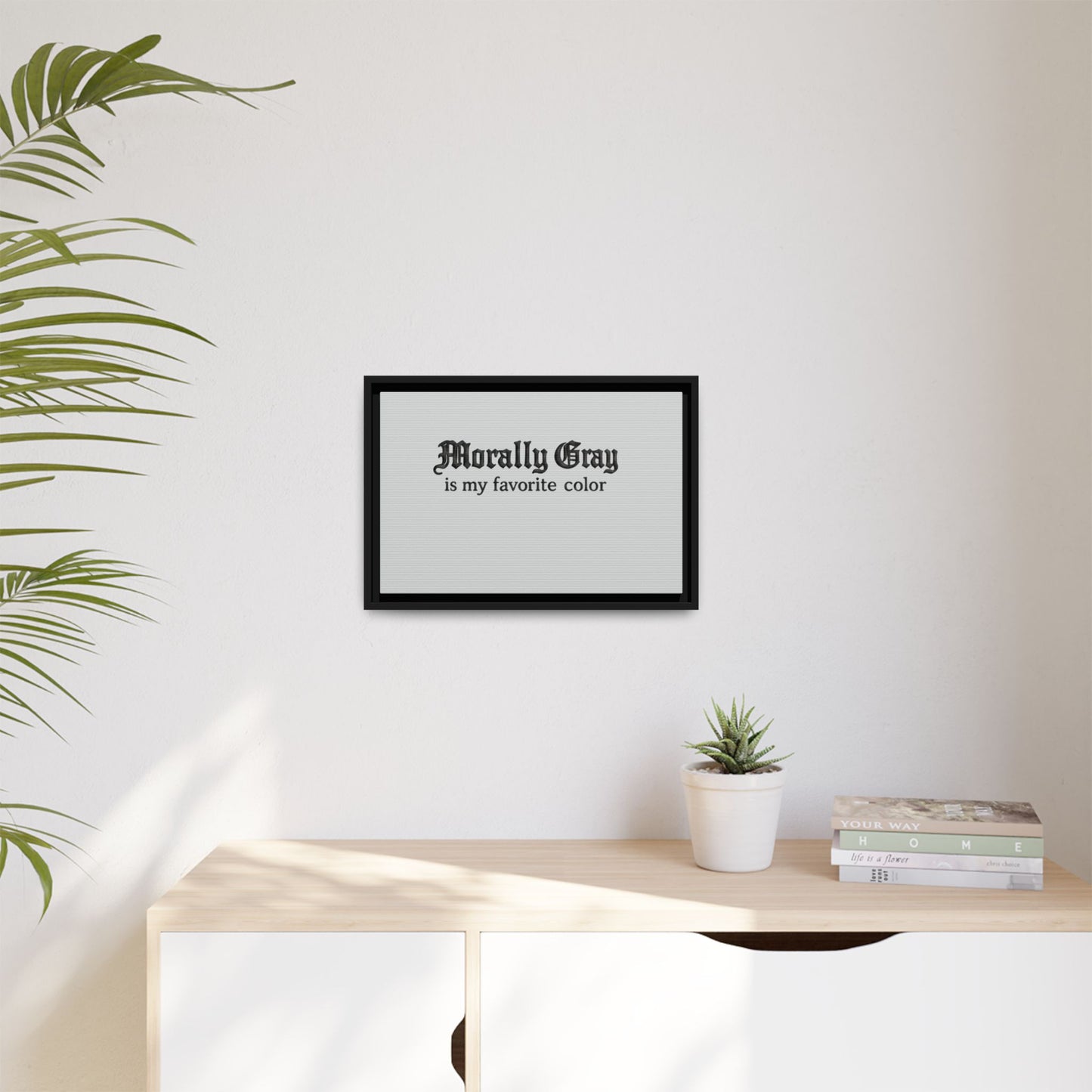 Framed wall art with the phrase "Morally Gray is my favorite color" in black text above a light wooden dresser with a small plant and stack of books in a minimalist room featuring the Unknown Product.