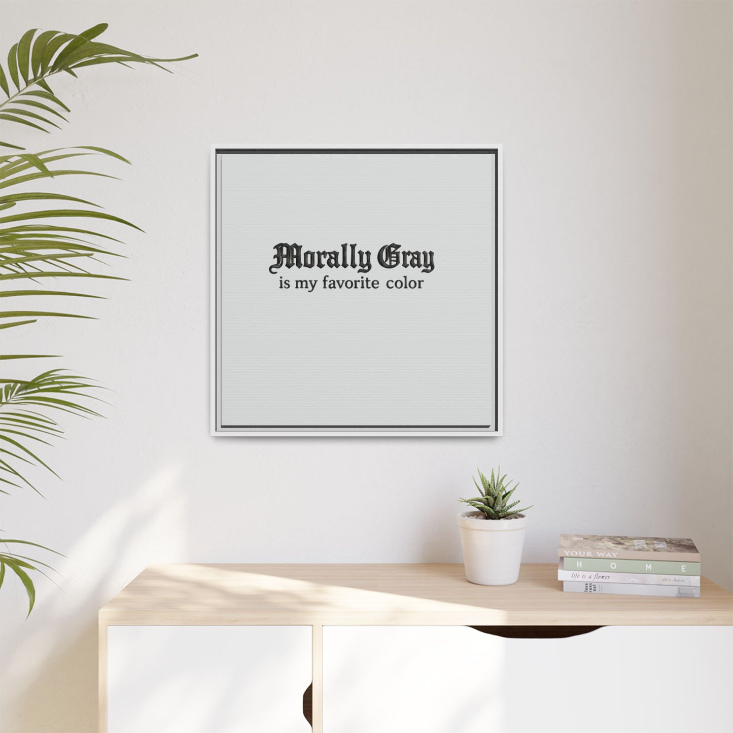 Minimalist wall art with the phrase "Morally Gray is my favorite color" in gothic font above a wooden surface, featuring a small potted plant and stacked books, showcasing Unknown Product.