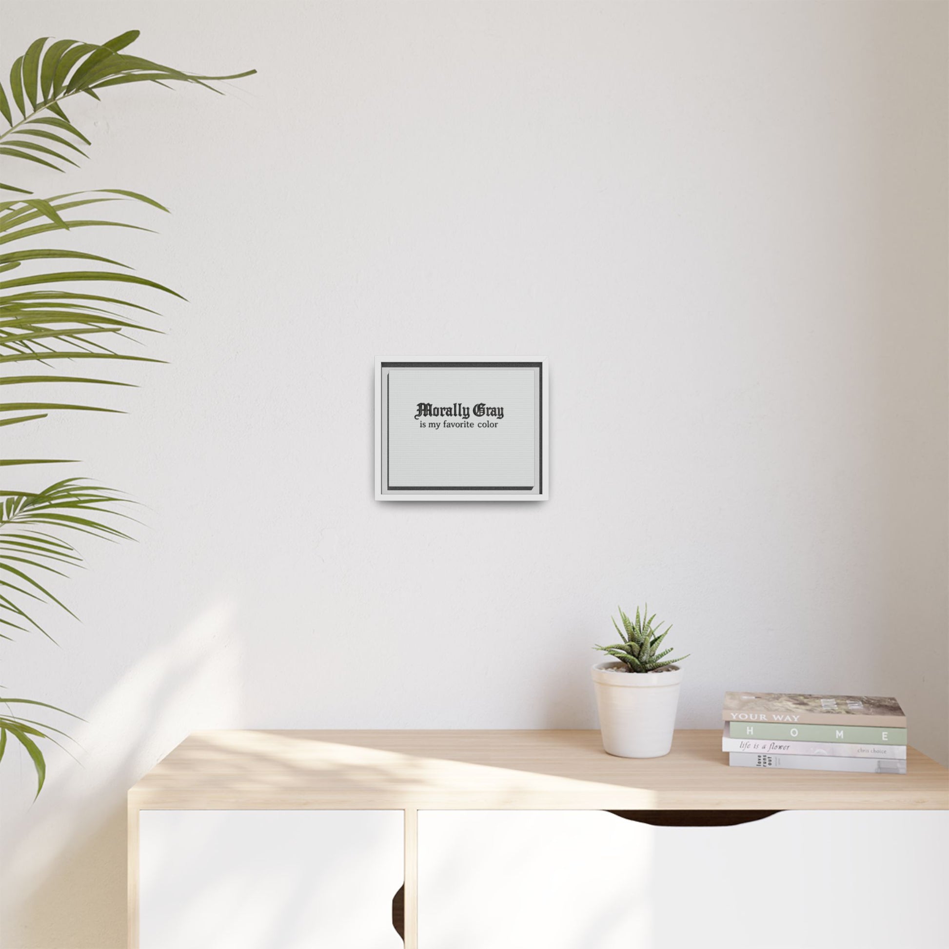 Minimalist home interior with a potted plant, stacked books, and a framed print on the wall reading "Morally Gray - a/my favorite color" featuring the Unknown Product.